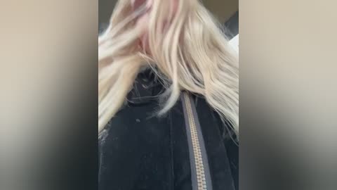 Video of a person with long, platinum blonde hair partially covering their face, wearing a black jacket with a visible zipper. The background is blurred, suggesting an indoor setting with neutral tones.