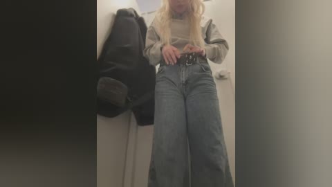 A video of a blonde woman with long hair, wearing a grey sweater and high-waisted, wide-leg jeans, adjusting her belt, standing in a hallway with a black backpack and a white door in the background.