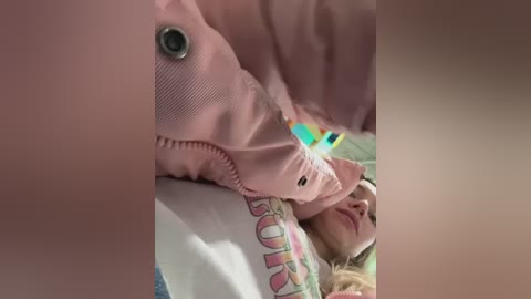 Video of a young woman with fair skin and blonde hair, lying on a bed. She wears a pink jacket and a colorful, patterned pillowcase. The image is slightly blurred, focusing on her face and the bed's soft texture.