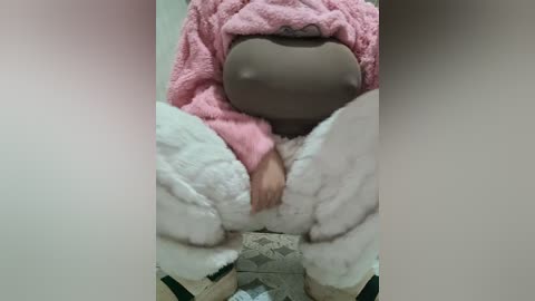 Media: A video of a person wearing a pink plushie onesie with a brown mask covering their face, sitting on a tiled floor. Their legs are spread apart, revealing white plushie pants.