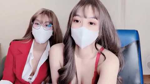 Media: Video of two East Asian women with long brown hair, wearing white face masks, red blazers, and glasses, sitting on blue chairs against a beige wall.