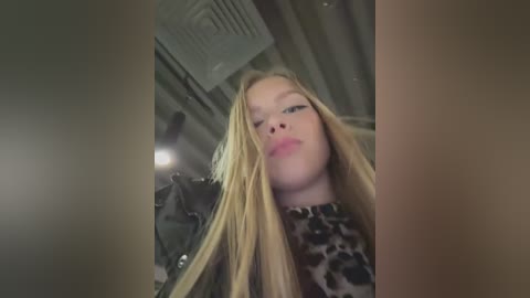 A video of a young woman with long, straight blonde hair, wearing a leopard print top, taken from a low angle, showing her from the shoulders up. Background includes a ceiling vent and blurred objects.