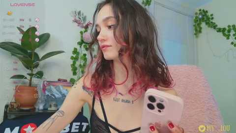 Media: A young woman with long, wavy black hair with pink highlights, tattoos, and a black bra takes a selfie in a cozy room with a calendar, plants, and pastel walls.