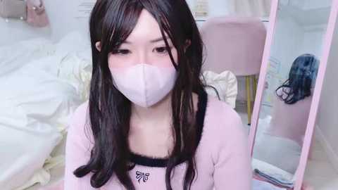 Media: Video of a young East Asian woman with long black hair, fair skin, and wearing a pink mask, pink top, and black bra, seated in a softly lit, pastel bedroom.