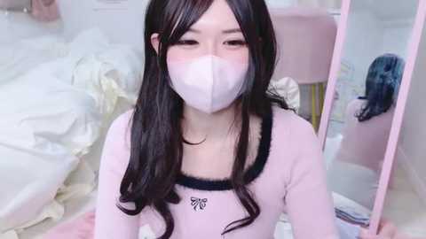 Media: Video of an East Asian woman with long, wavy black hair, wearing a light pink, long-sleeved top with a butterfly logo, a white face mask, and a soft pink room with a bed and mirror in the background.