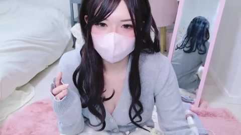 Media: A video of an East Asian woman with long, wavy black hair, wearing a white face mask, gray cardigan, and a low-cut top, kneeling on a pink rug, with a mirror reflecting her face in a bedroom.