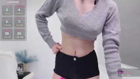 Media: Video of a slim, light-skinned woman with medium breasts, wearing a gray, ribbed cropped sweater and black shorts, standing indoors. Background features a calendar and blurred decor.