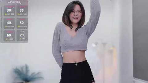 Media: Video of a smiling Asian woman with shoulder-length black hair, wearing a light grey cropped top and black high-waisted pants, standing in a minimalist, brightly lit room with a temperature display on the wall.