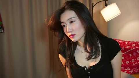 Video of an East Asian woman with long black hair and fair skin, wearing a black blouse with a plunging neckline, in a dimly-lit bedroom with a red-patterned pillow and a lamp.