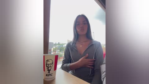 Media: Video of a young woman with medium brown skin, long black hair, wearing a gray hoodie, standing in a fast-food restaurant, holding a KFC cup.