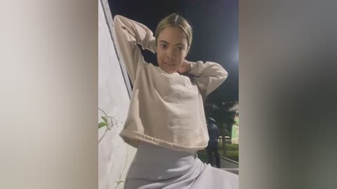 Media: Video of a blonde woman with light skin, wearing a loose, light pink sweater and a high-waisted light grey skirt, leaning against a white wall outdoors at night.