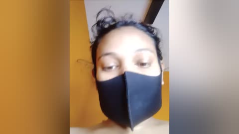 Media: Video of a person with short curly black hair, wearing a black face mask and a dark top, standing against a mustard-yellow wall.