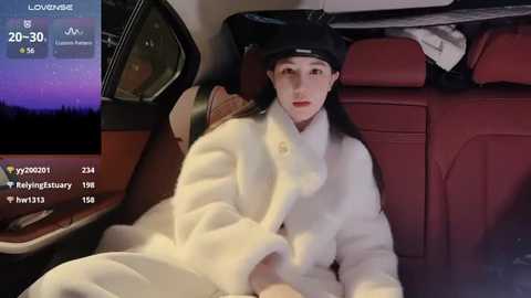 Media: A video shows a young woman with long black hair and fair skin, wearing a black hat and white robe, reclining on a red leather seat in a car.