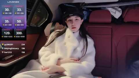 Media: Video of a young Asian woman with long black hair, wearing a white fur coat and black cap, sitting in a car's backseat, surrounded by plush red seats.