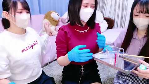 Media: Video of three Asian women in face masks, wearing blue gloves, in a soft, pastel room. One holds a tray, another a cup, and the third a box. Background features a plush teddy bear.