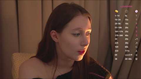 Video of a young Asian woman with straight black hair, wearing a black off-shoulder top, sitting indoors with dim lighting. Background shows a beige curtain and a yellow cushion.