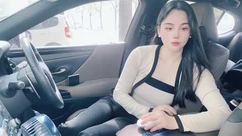 Media: A video of an Asian woman with long black hair, wearing a beige cardigan and black pants, driving a car with a beige interior and a white car visible through the window.