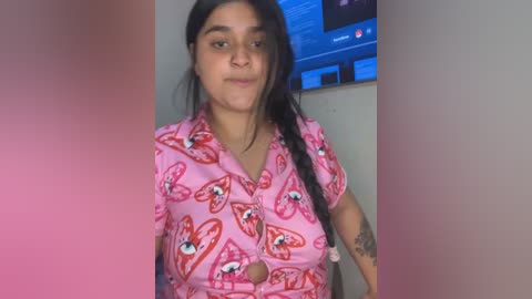 Media: Video of a young woman with medium skin tone, dark hair, and a curvy physique, wearing a pink shirt with butterfly patterns, smiling softly, in a room with a TV screen displaying text.