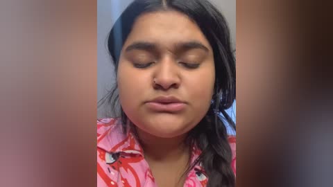 Media: Video of a South Asian woman with medium brown skin and long black hair, wearing a pink floral blouse, eyes closed, appearing contemplative.