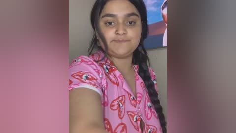 Media: Video of a young woman with long dark hair, wearing a pink shirt with red heart patterns, standing indoors. The background features a blurred painting of a woman.