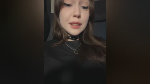 Media: Video of a young woman with fair skin, brown hair, wearing a black choker necklace and a black top, looking down with a neutral expression.