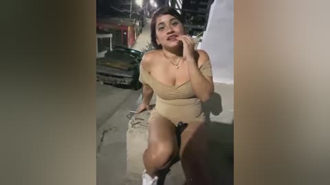 Media: Video of a Latina woman with light brown skin, wearing a beige off-shoulder romper, sitting on a curb in a dark, industrial setting, holding a bag and looking contemplative.