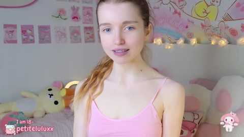 Media: A young, fair-skinned girl with long, blonde hair in pigtails, wearing a pink spaghetti-strap top, sits in a pink-themed bedroom adorned with plush toys, pastel decorations, and fairy lights.