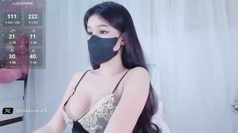 Media: Video of an Asian woman with long black hair, wearing a black face mask, black spaghetti strap top, and sheer lace bralette, in a bright, minimalist room.