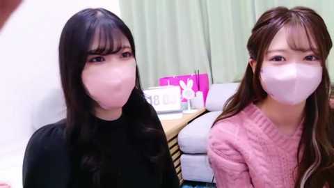 Media: Video of two East Asian women with long dark hair, wearing face masks, seated indoors. One wears a black top, the other a pink sweater. Background includes a white couch, a pink backpack, and a white table.