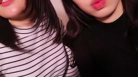 Media: Video of two Asian women with long black hair, one wearing a black top, the other a white striped top, both with full lips, close-up.