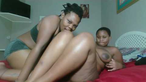 Media: Video of two African-American women in a bedroom; one topless with large breasts, the other in a gray bra and shorts, both smiling.