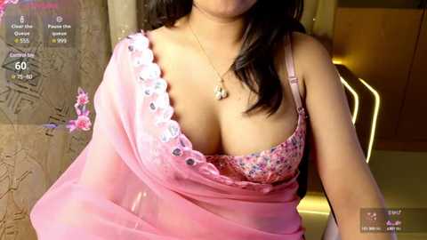 Media: Video of a South Asian woman with medium skin tone, wearing a pink saree with floral pattern, revealing a pink bra underneath. She has long, dark hair and wears a gold necklace. Background shows a warm, dimly-lit room.