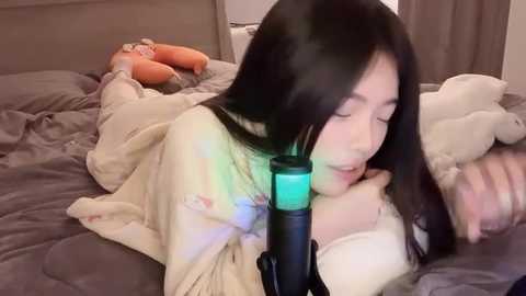 Media: Video of a young Asian woman with long black hair, wearing a beige robe, lying on a bed with gray sheets, illuminated by a green light, surrounded by plush toys.