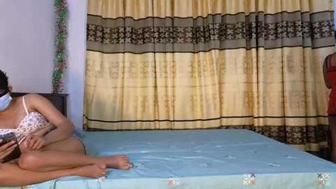 Media: Video of a woman in leopard-print lingerie, sitting on a bed with light blue sheets, wearing a face mask, in a room with beige and brown patterned curtains.