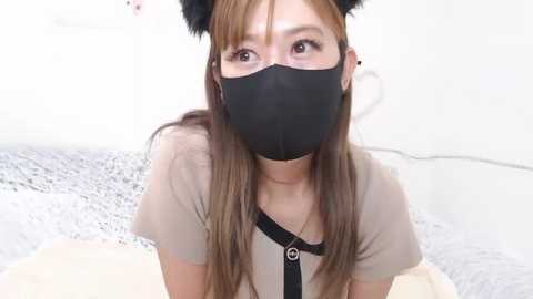 Media: Video of an Asian woman with long brown hair, wearing black cat ear headband, black mask, and beige top, in a minimalist, white-walled room.