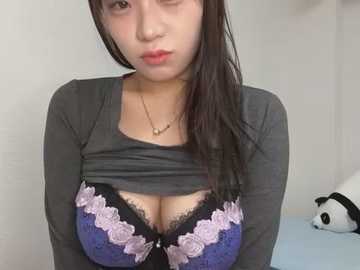 Media: Video of an Asian woman with long black hair, wearing a black top pulled up to reveal a purple lace bra with pink flowers, and a gold necklace. Background includes a white wall and a stuffed panda.