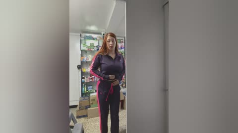 Media: A video of a woman with light skin and red hair, dressed in a purple tracksuit, standing in a cluttered room with shelves full of various items.