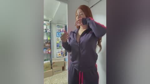 Video of a woman with long, auburn hair, wearing a dark purple tracksuit with red stripes, in a supermarket aisle. She appears to be sneezing into her elbow, surrounded by shelves of colorful products.