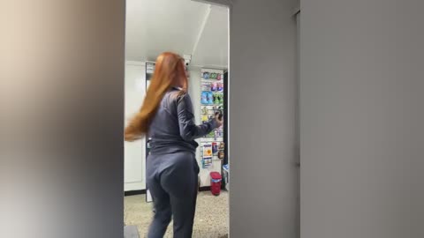 Media: Video of a woman with long, straight red hair, wearing a navy blue jumpsuit, walking out of a brightly lit store with colorful magazines on a shelf in the background.