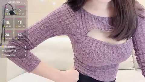 Video of a woman with fair skin and straight, dark brown hair, wearing a purple ribbed sweater with a cutout design, standing indoors against a beige wall.