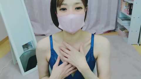 Media: Video of an East Asian woman with short dark hair, wearing a blue sleeveless dress, a face mask, and holding her chest, in a bedroom with light purple curtains and white furniture.