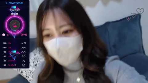 Media: A video of an Asian woman with long dark hair, wearing a white face mask, gray sweater, and lying on a blue cushion. A digital overlay shows her \"Lovesense\" stats, including heart rate and temperature.