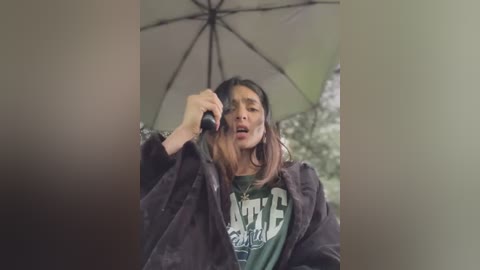 Media: Video of a young woman with shoulder-length hair, wearing a green graphic t-shirt and a dark jacket, holding a phone under a green umbrella in a dimly lit, cluttered room.