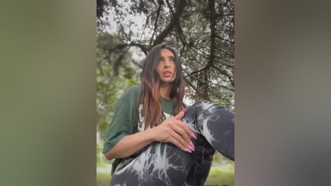 Media: Video of a young woman with long, straight brown hair, wearing a green T-shirt and black pants with white paint splatter, sitting under a tree in a park.