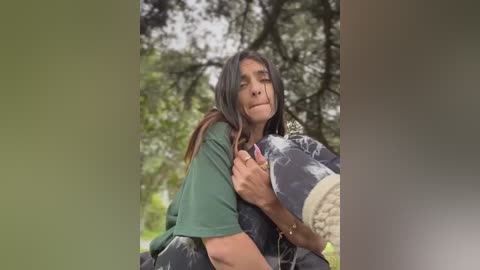 Media: Video of a solemn man with long hair, wearing a green t-shirt, hugging a woman in a grey jacket with a beige fur collar, set against a blurred, green forest background.