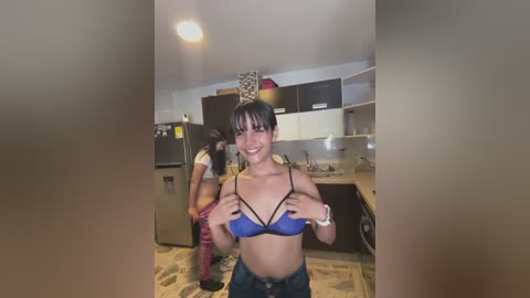 Media: A video of two women in a kitchen. One woman with dark hair, wearing a blue bra, smiles. The other, with long hair, is bending over a refrigerator. The kitchen has dark cabinets and a tiled floor.