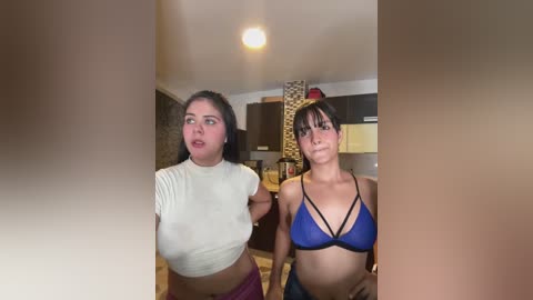 Media: Video of two young women with different body types, one in a tight white crop top and the other in a blue bikini top, in a modern kitchen.