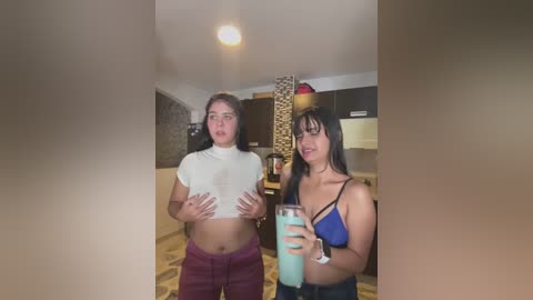 Media: Video of two young women in a modern kitchen: one with a white crop top, the other in a blue spaghetti strap top, both holding drinks.