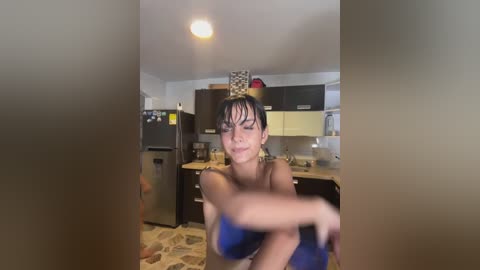 Media: A video shows a woman with wet hair, wearing a yellow hat, sitting topless in a kitchen with stone-patterned flooring.