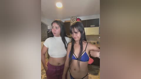 Media: Video of two women in a modern kitchen; one in a white crop top and maroon pants, the other in a blue bikini.